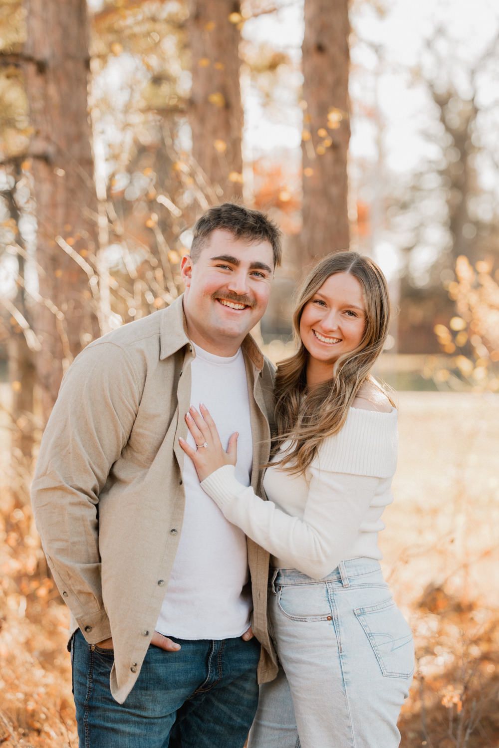 A couple embraces and laughs together in a forest setting during golden hour wearing coordinated beige and white outfits.