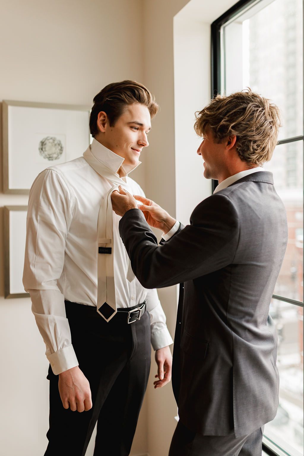 Two people in formalwear adjusting their attire while standing by a window in a light-filled room.