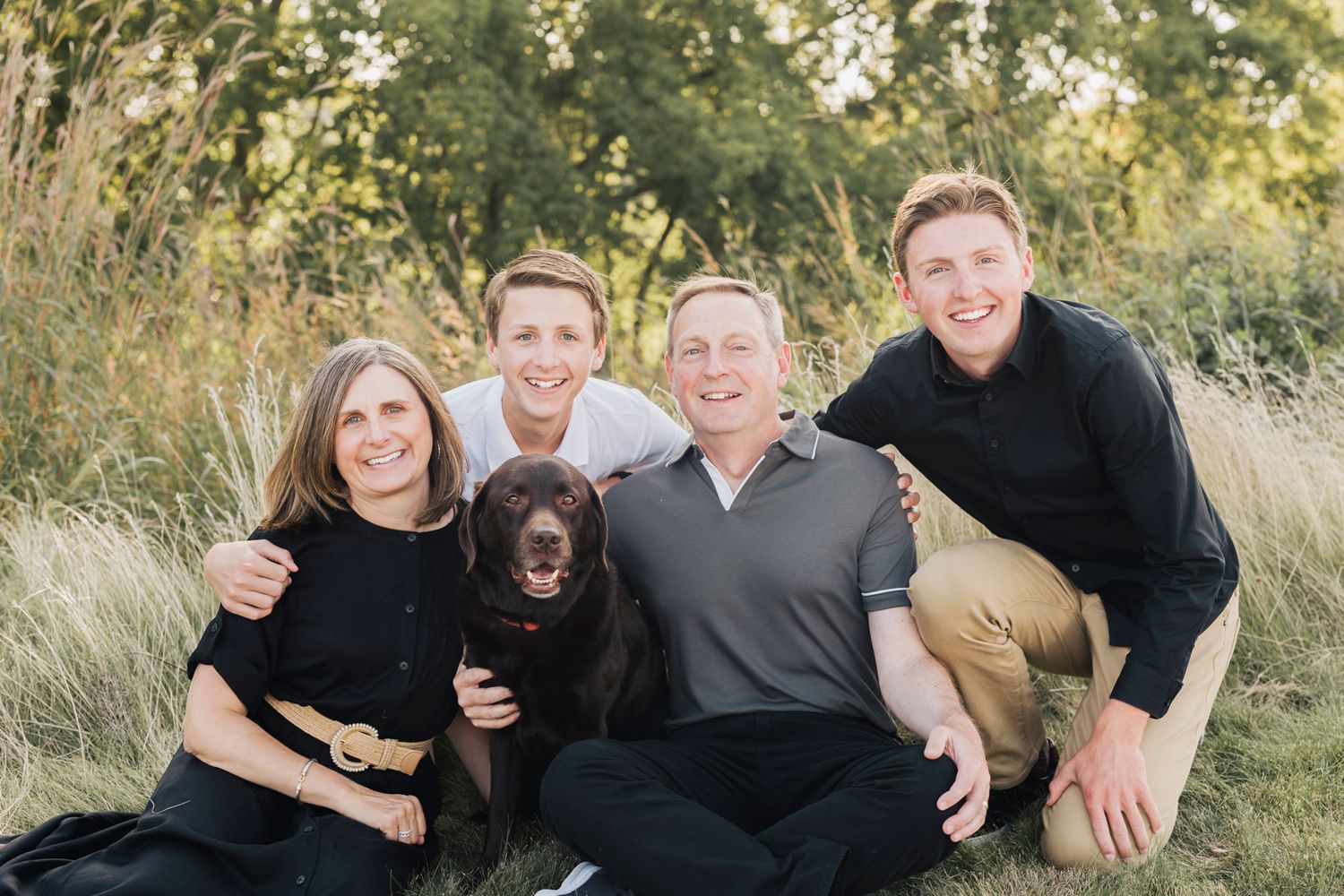 Family group relaxes on grass with their dog during outdoor photo session.
