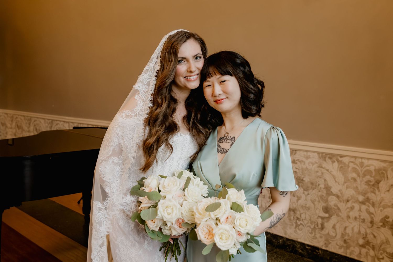 Bride in lace veil holding white bouquet poses with bridesmaid in sage green dress at wedding.