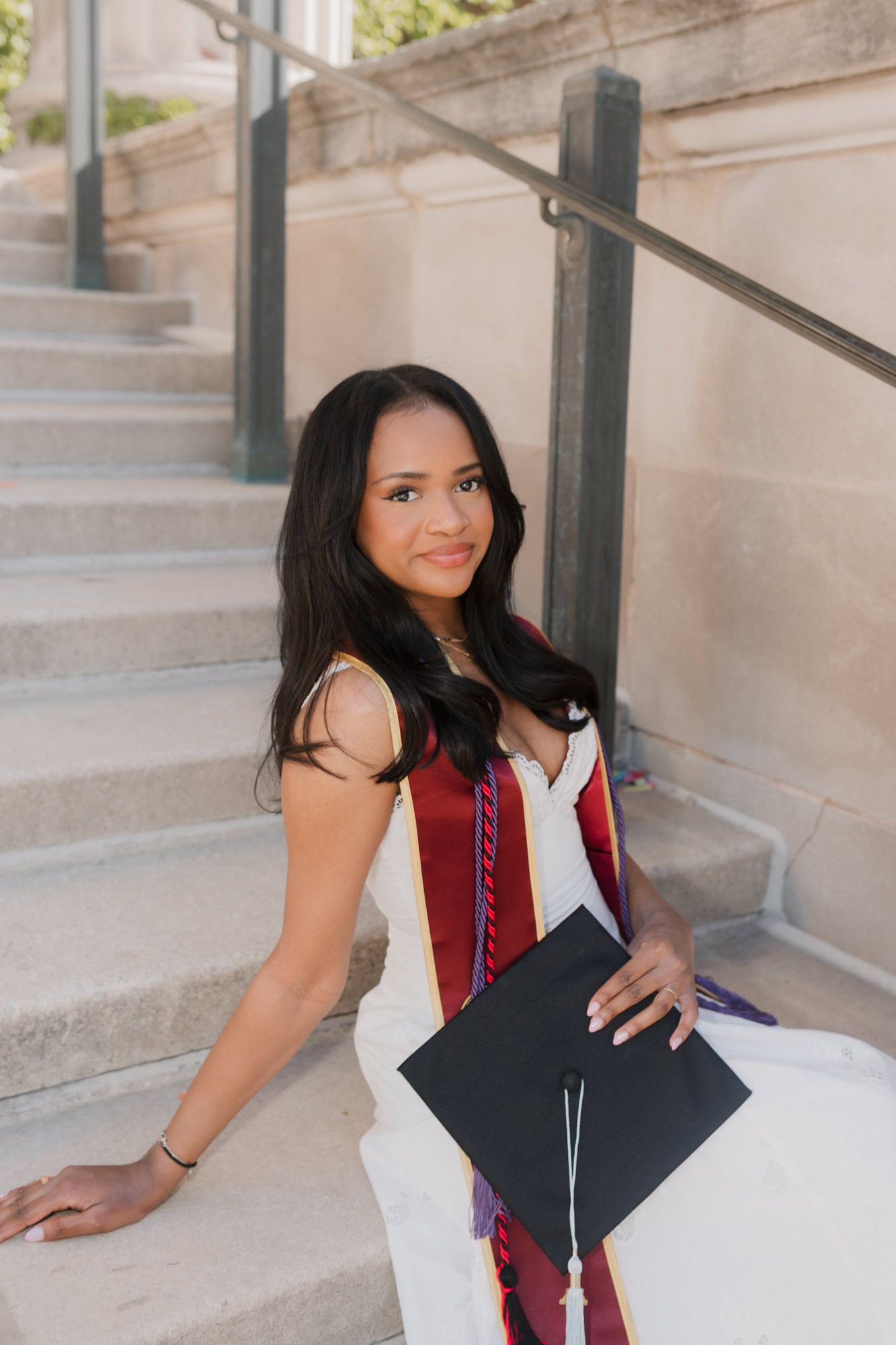 Young woman in white dress holding graduation cap on stone steps, wearing red sash, celebrating academic achievement.