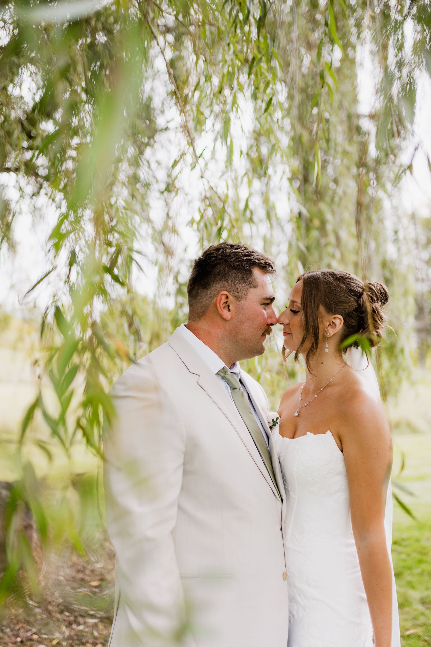 Bride and groom sharing romantic nose-to-nose moment under weeping willow branches on wedding day.