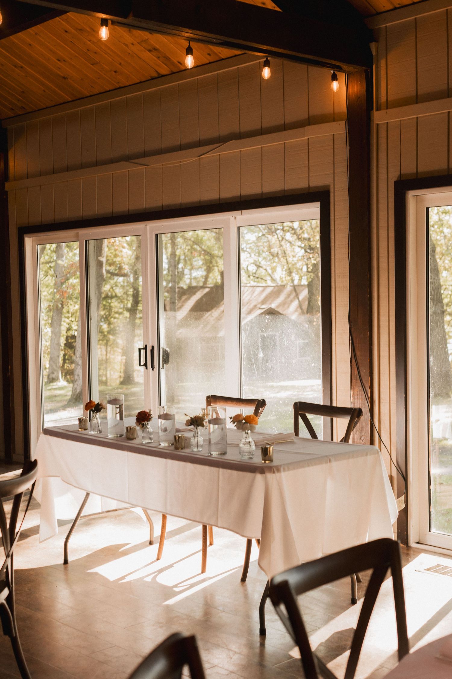 Elegant dining table with white tablecloth set against large windows in a rustic room with wooden ceiling and string lights.