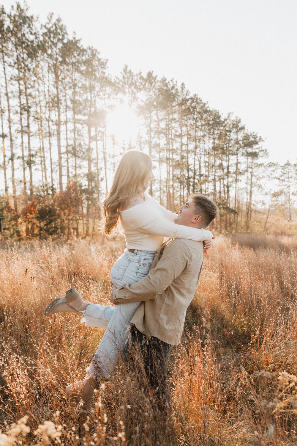 Romantic couple embraces in a golden field at sunset with tall pine trees silhouetted in the background.
