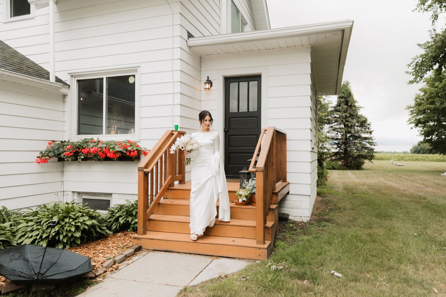 White farmhouse with wooden deck stairs and railings surrounded by green grass and landscaping.