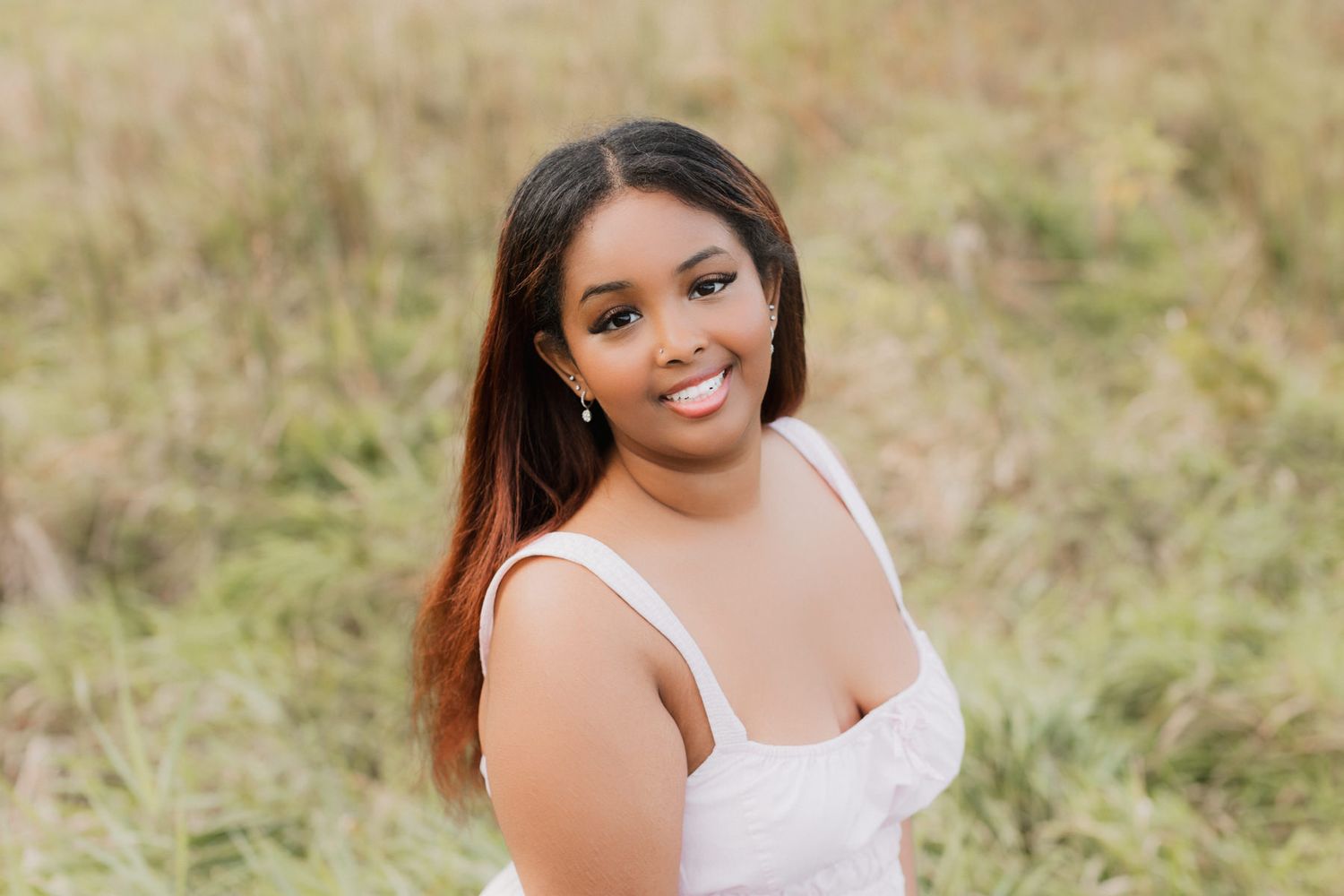 Smiling young woman in a white dress poses in a sunlit field of tall green grass.