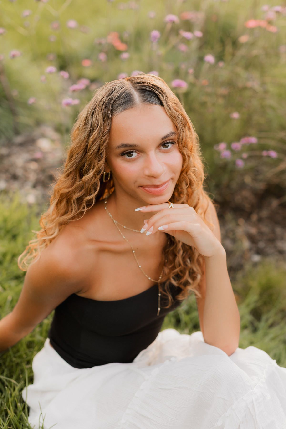 Young woman with curly auburn hair sits in a flower field wearing a black top and white skirt.