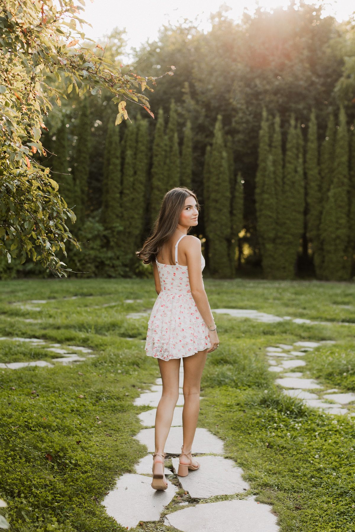 Young woman in floral mini dress walking stone garden path surrounded by lush green trees in golden sunlight.