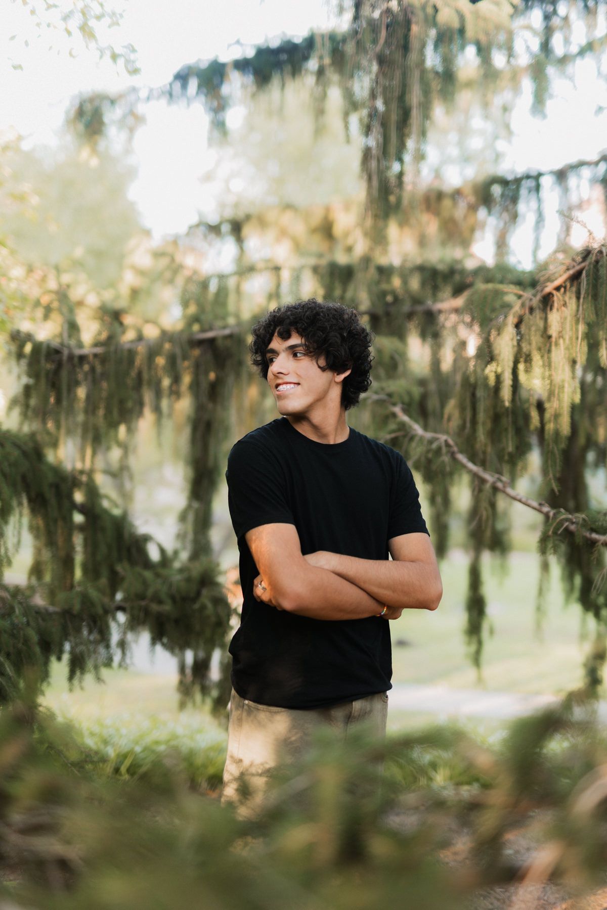 Young man in black t-shirt standing with arms crossed in a lush green forest setting.