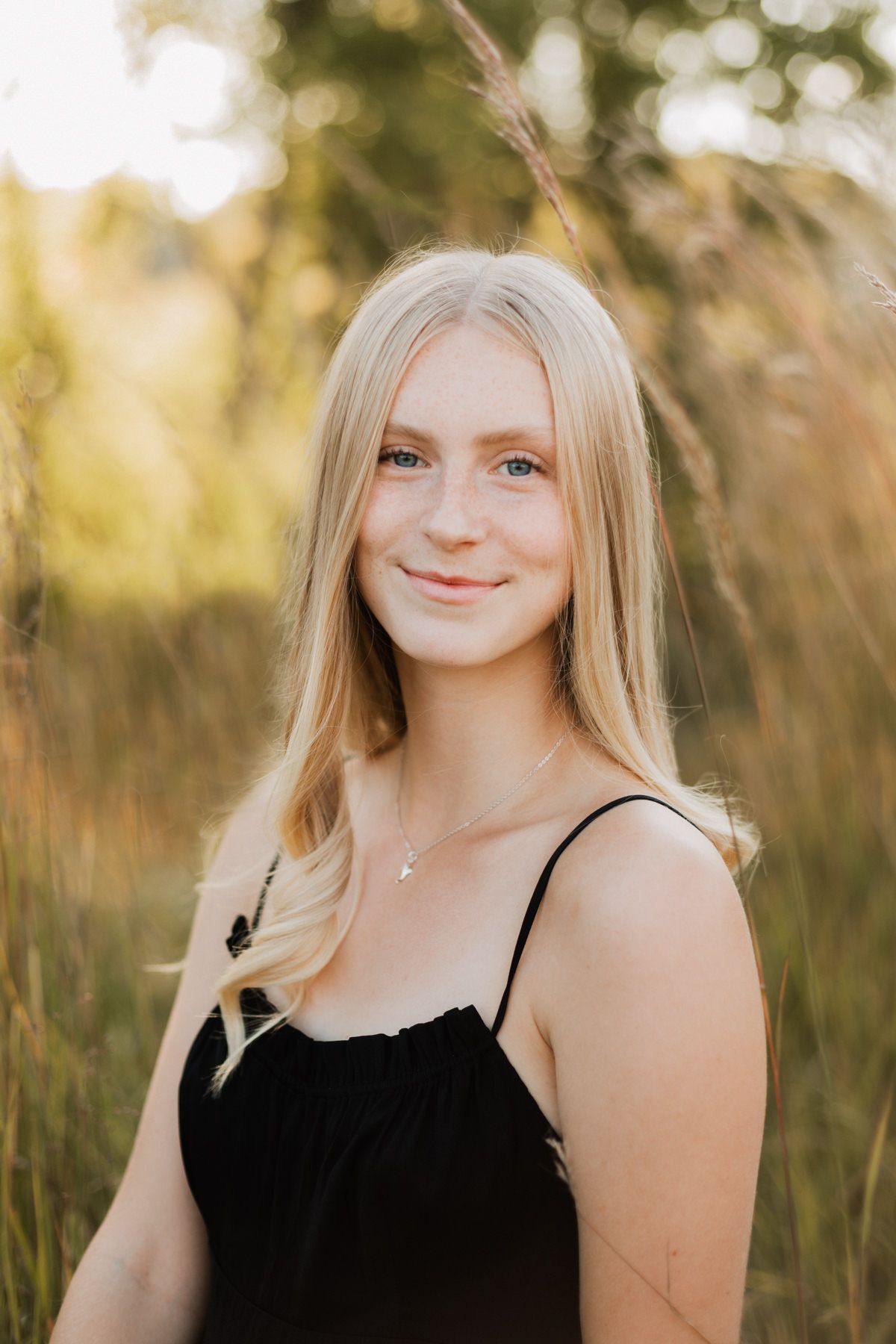 Smiling blonde girl in black spaghetti-strap dress wearing a delicate necklace, posing in a sunlit field.