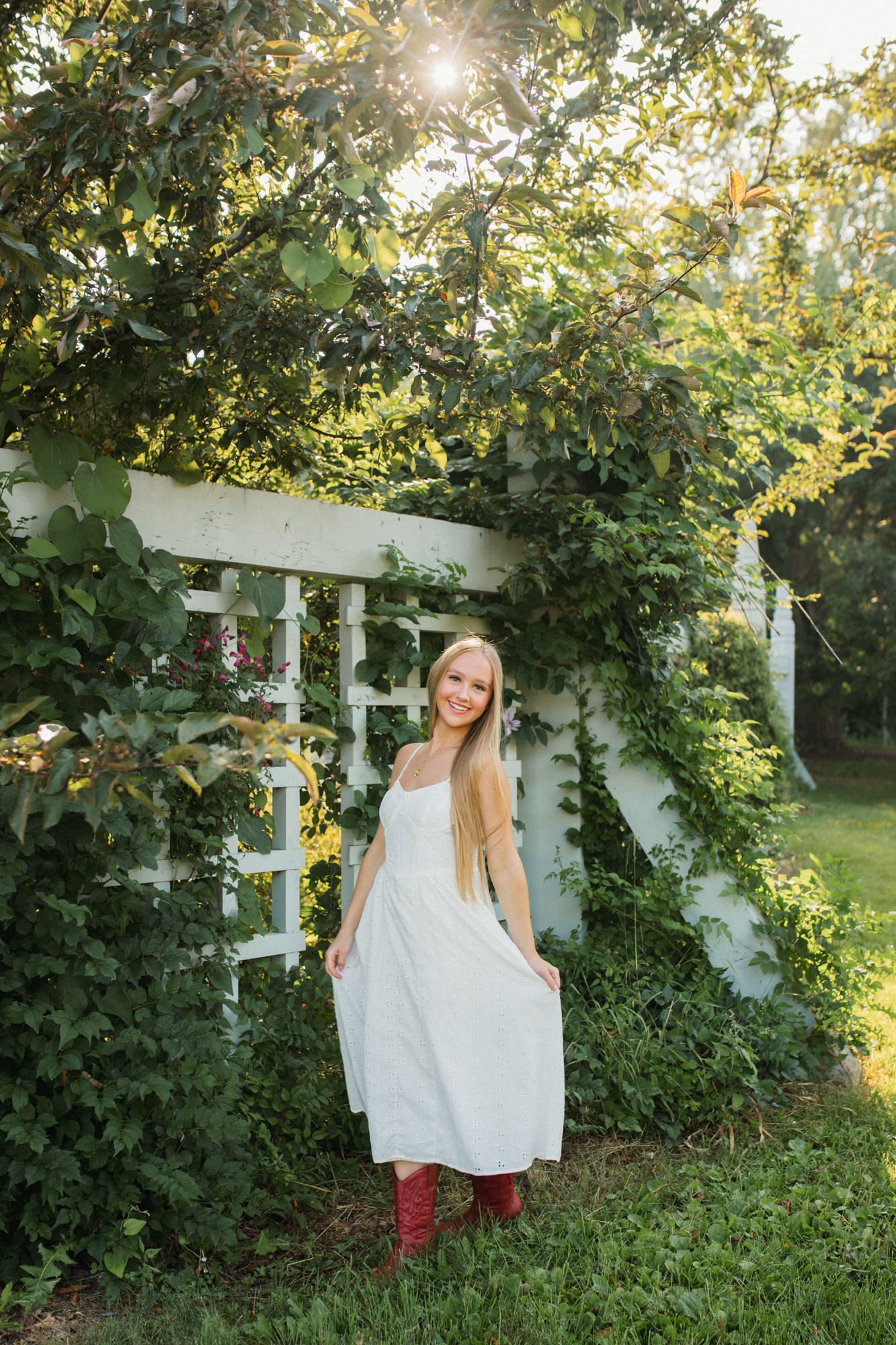 Young woman in white dress and red boots smiling in a lush green garden with white trellis and sunlight.