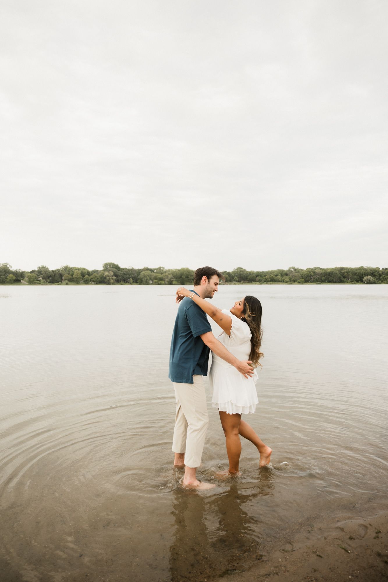 Couple embracing and dancing in shallow lake water, woman in white dress and man in blue shirt, with trees in background.