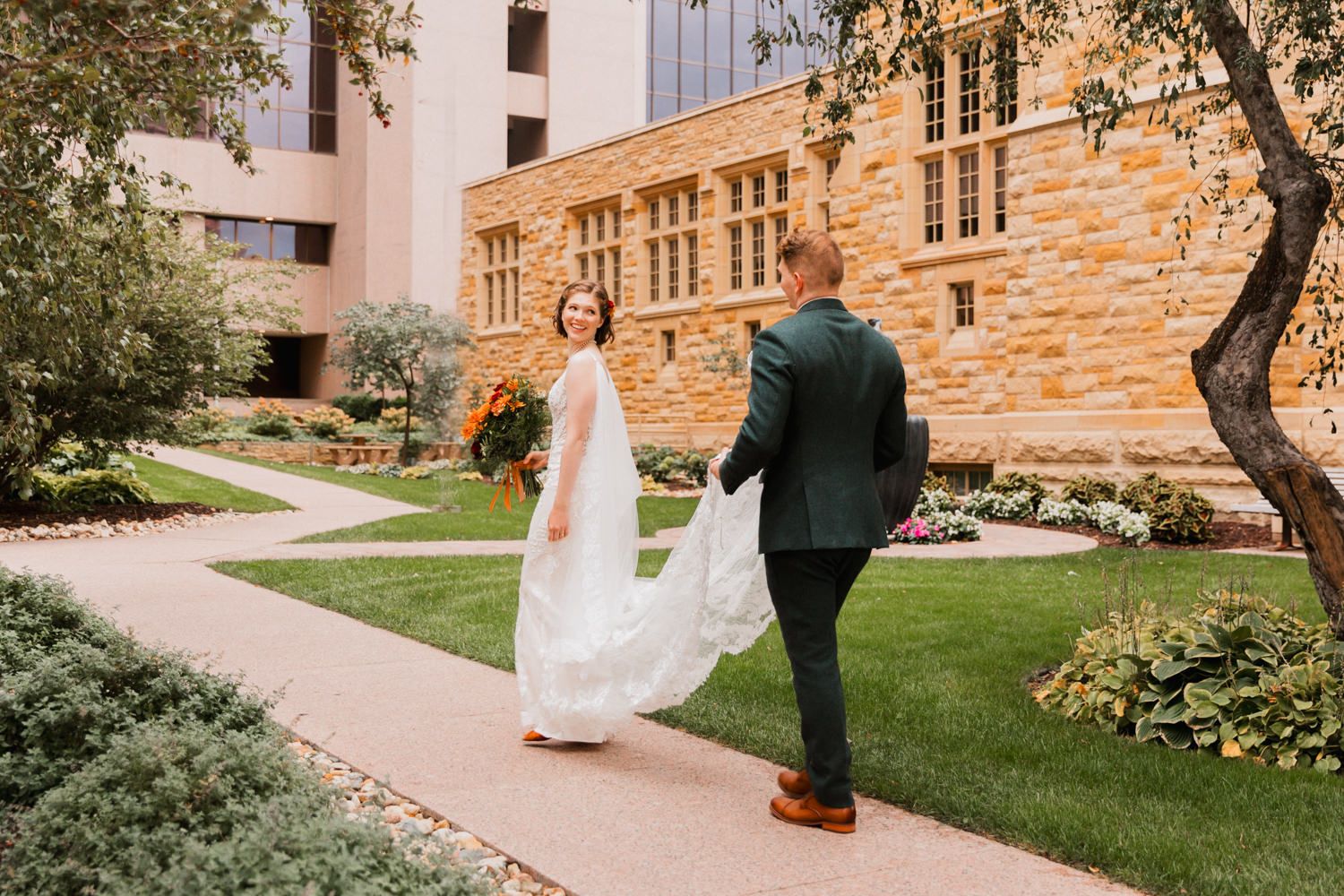 A couple walks along a stone building exterior with landscaped gardens on a sunny day.