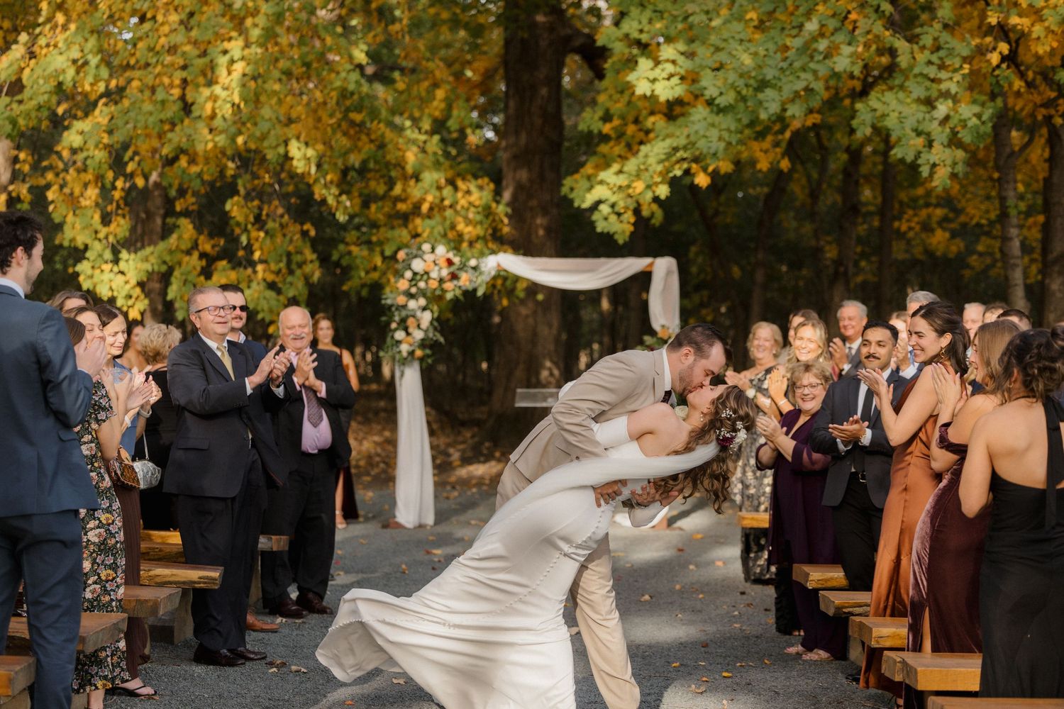 Outdoor autumn wedding ceremony as a groom dips his bride for a kiss while smiling guests applaud beneath golden fall foliage.