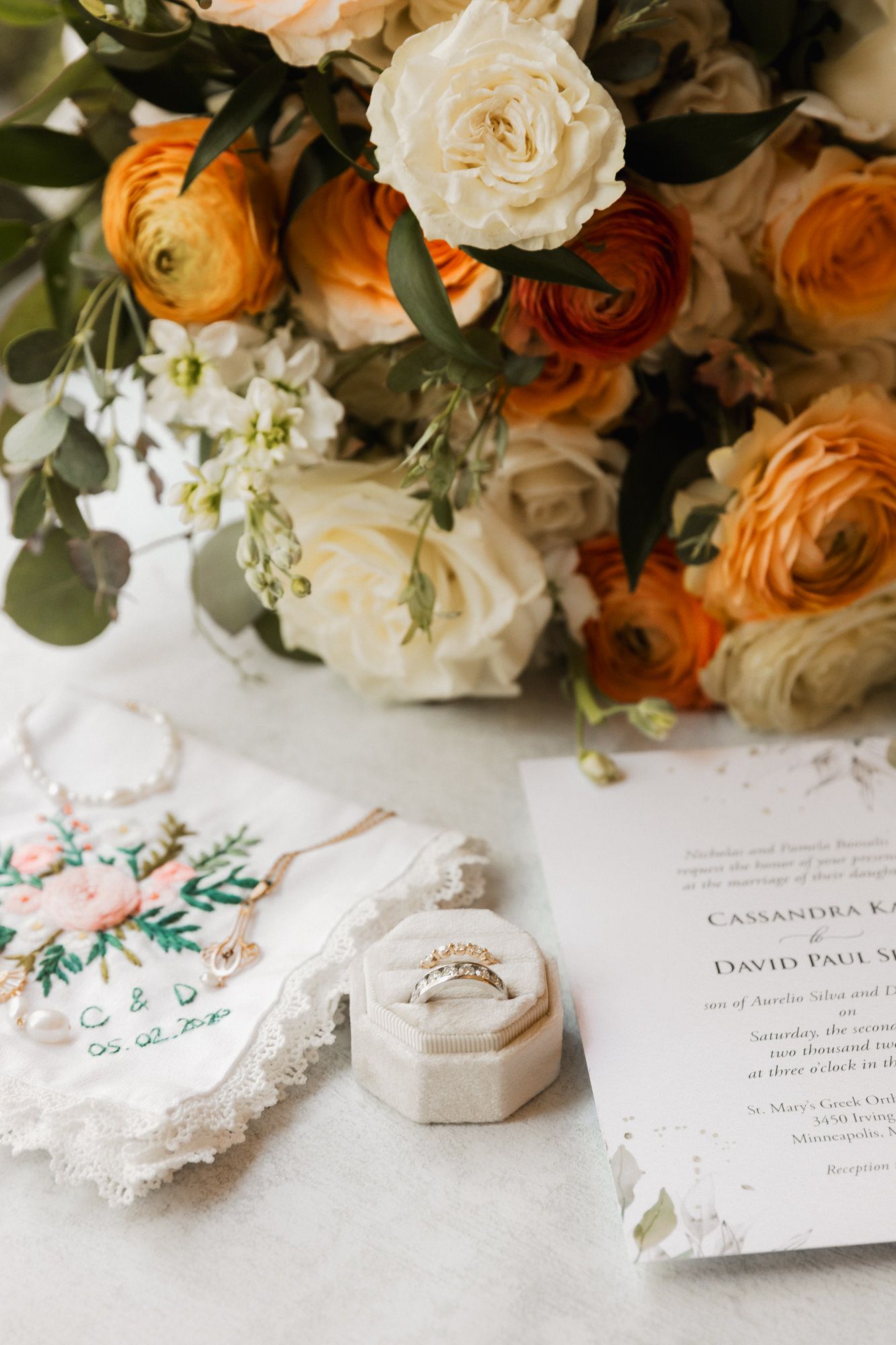 Wedding flat lay with orange and white floral bouquet, diamond ring in velvet box, and elegant invitation suite.