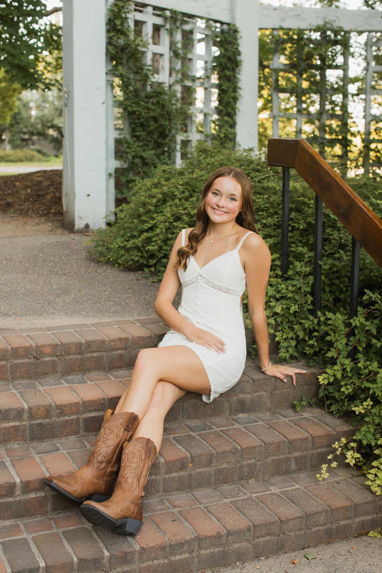 Young woman in white dress and cowboy boots sitting on brick steps near a garden pergola, smiling outdoors.