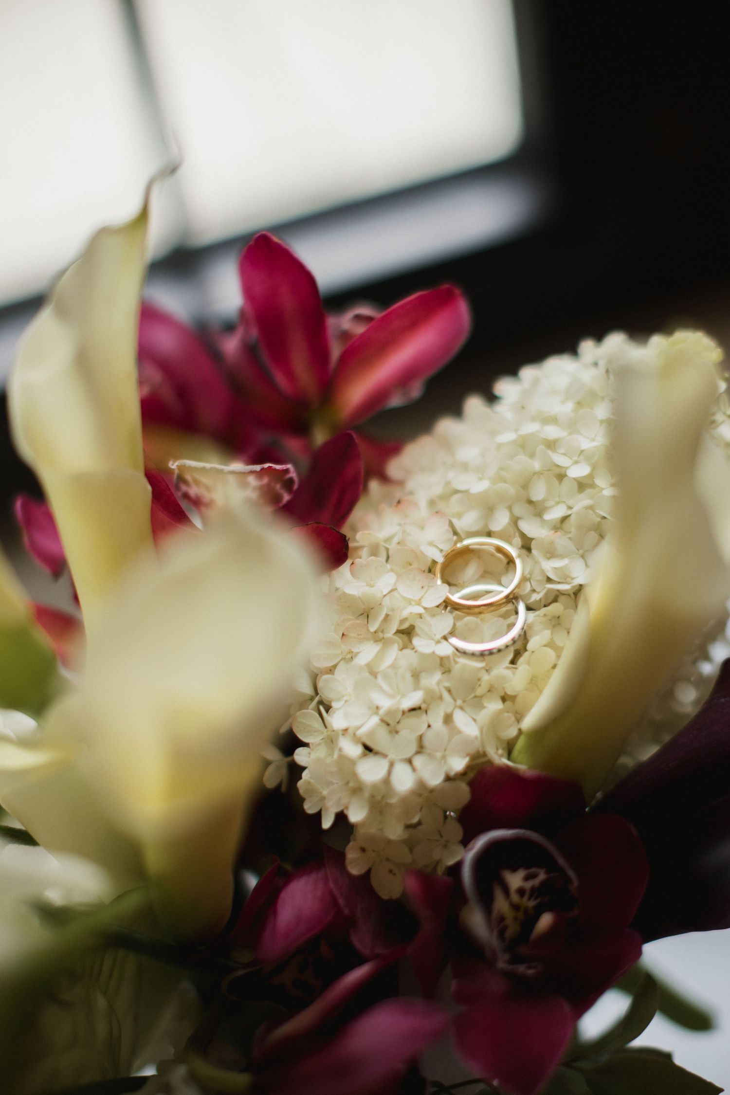 Two gold wedding rings rest on white lilac blooms surrounded by cream calla lilies and deep magenta orchids in a bridal bouquet.