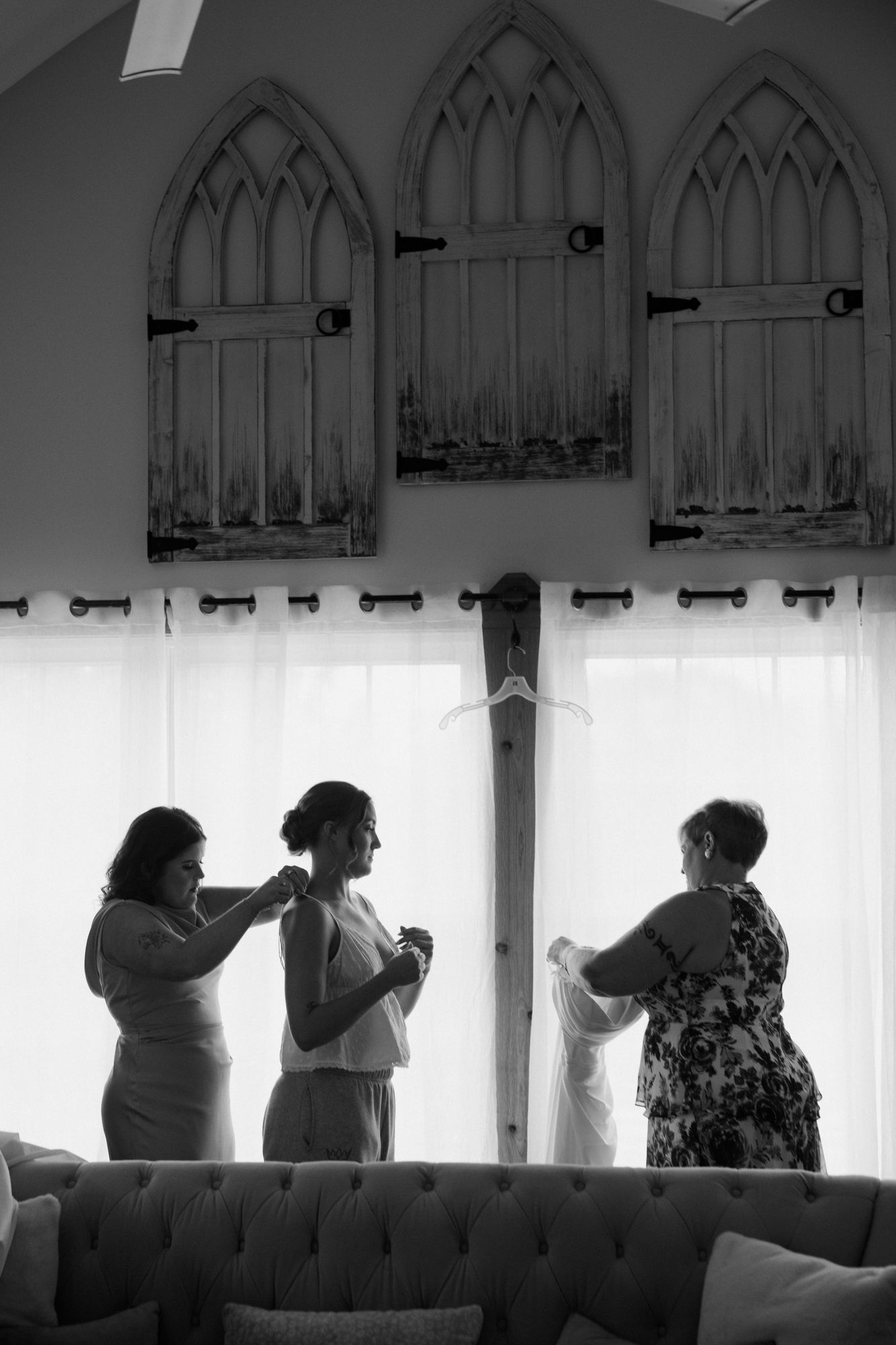 Black and white photo of bride being helped by bridesmaids before wedding ceremony in elegant room with gothic windows.