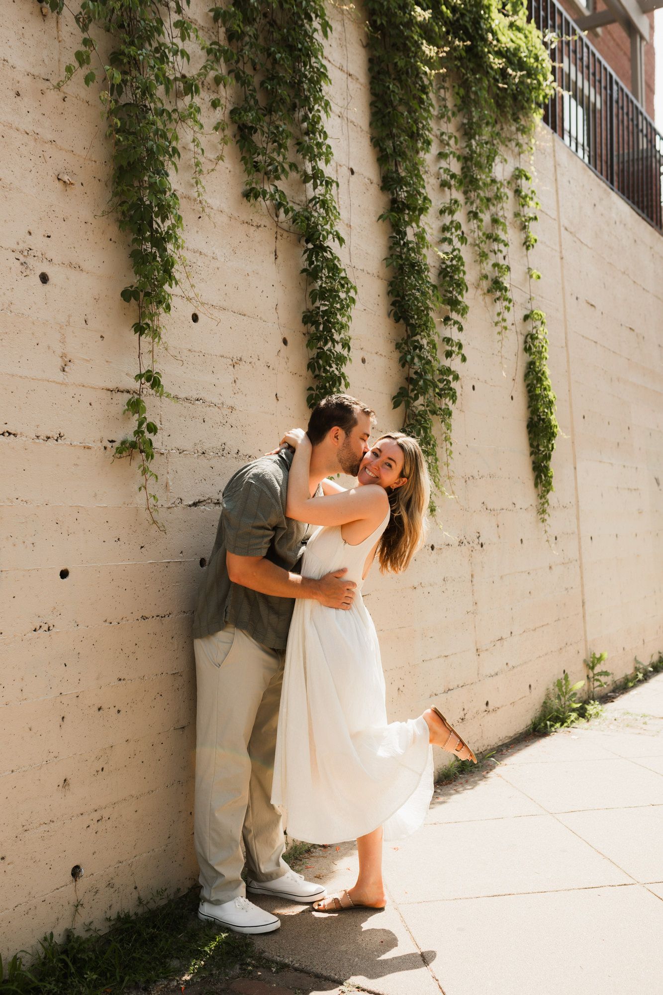 Couple embracing and kissing against a concrete wall with hanging ivy vines outdoors.
