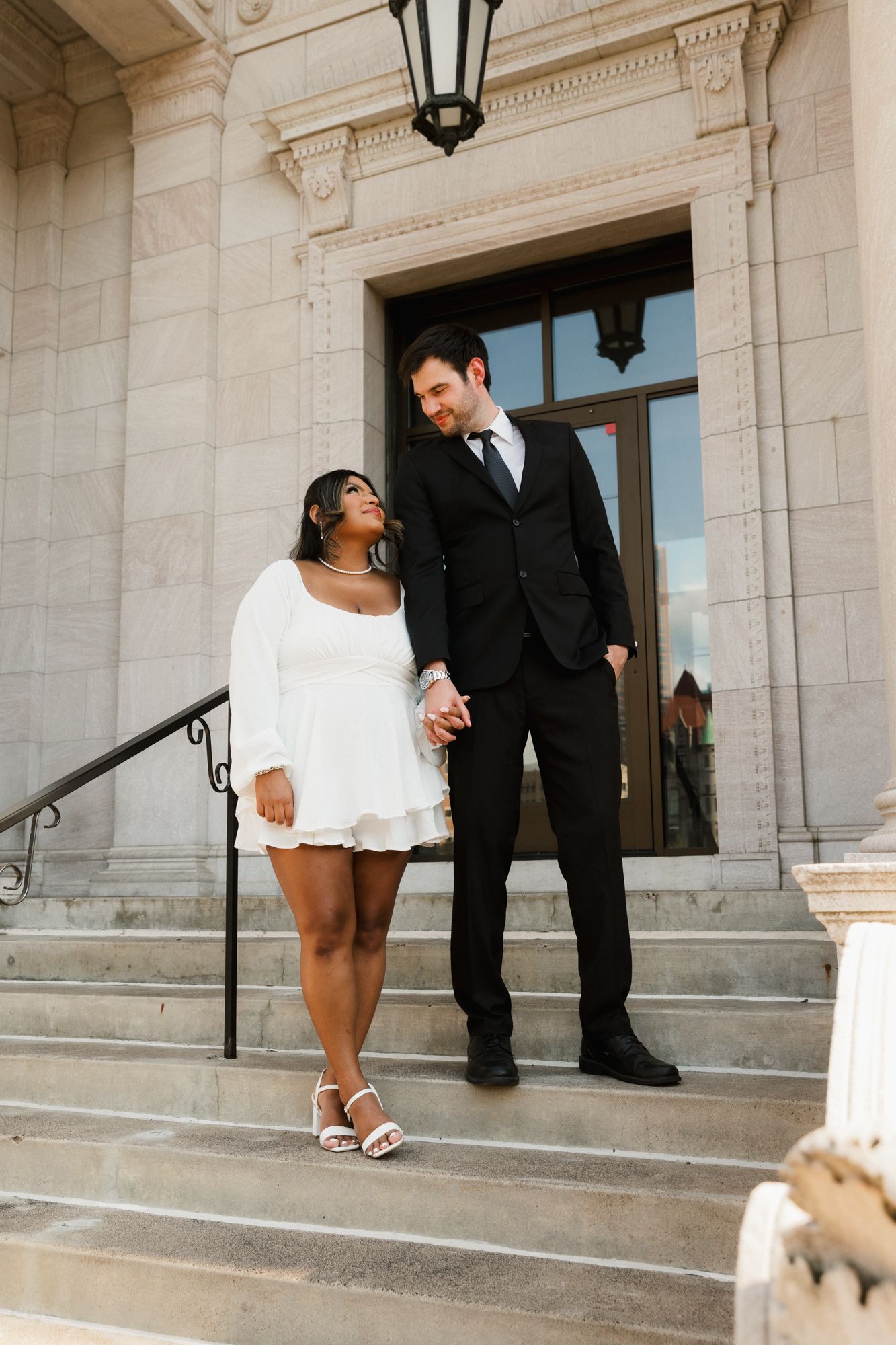 Couple in wedding attire holding hands on stone steps, woman in white mini dress and man in black suit.
