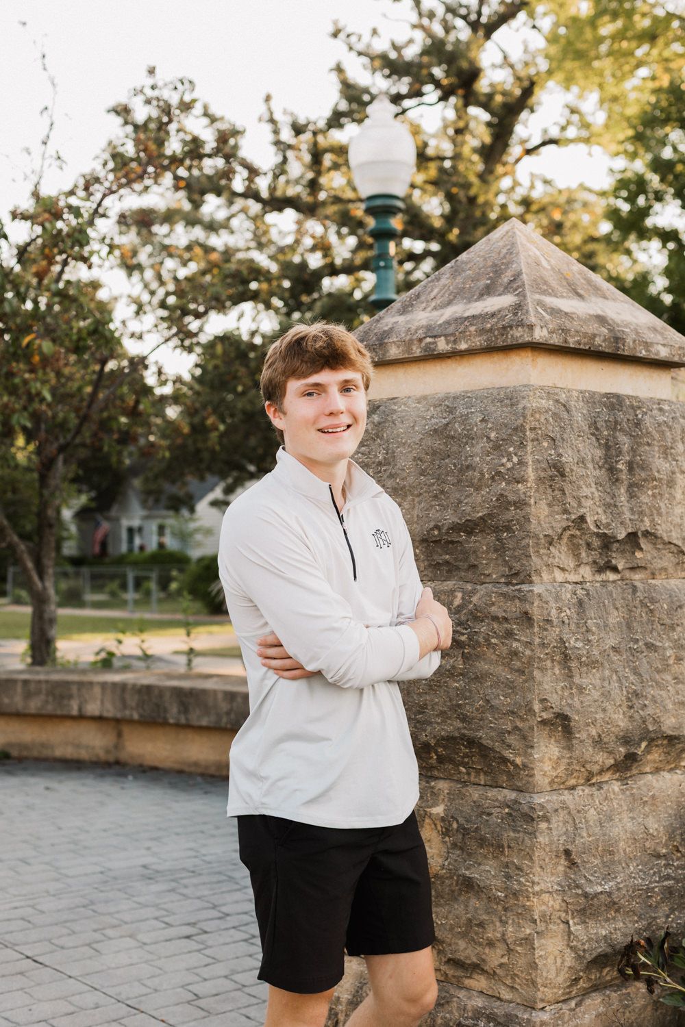 Person in white sweatshirt and black shorts poses by stone wall in outdoor setting with golden sunlight.