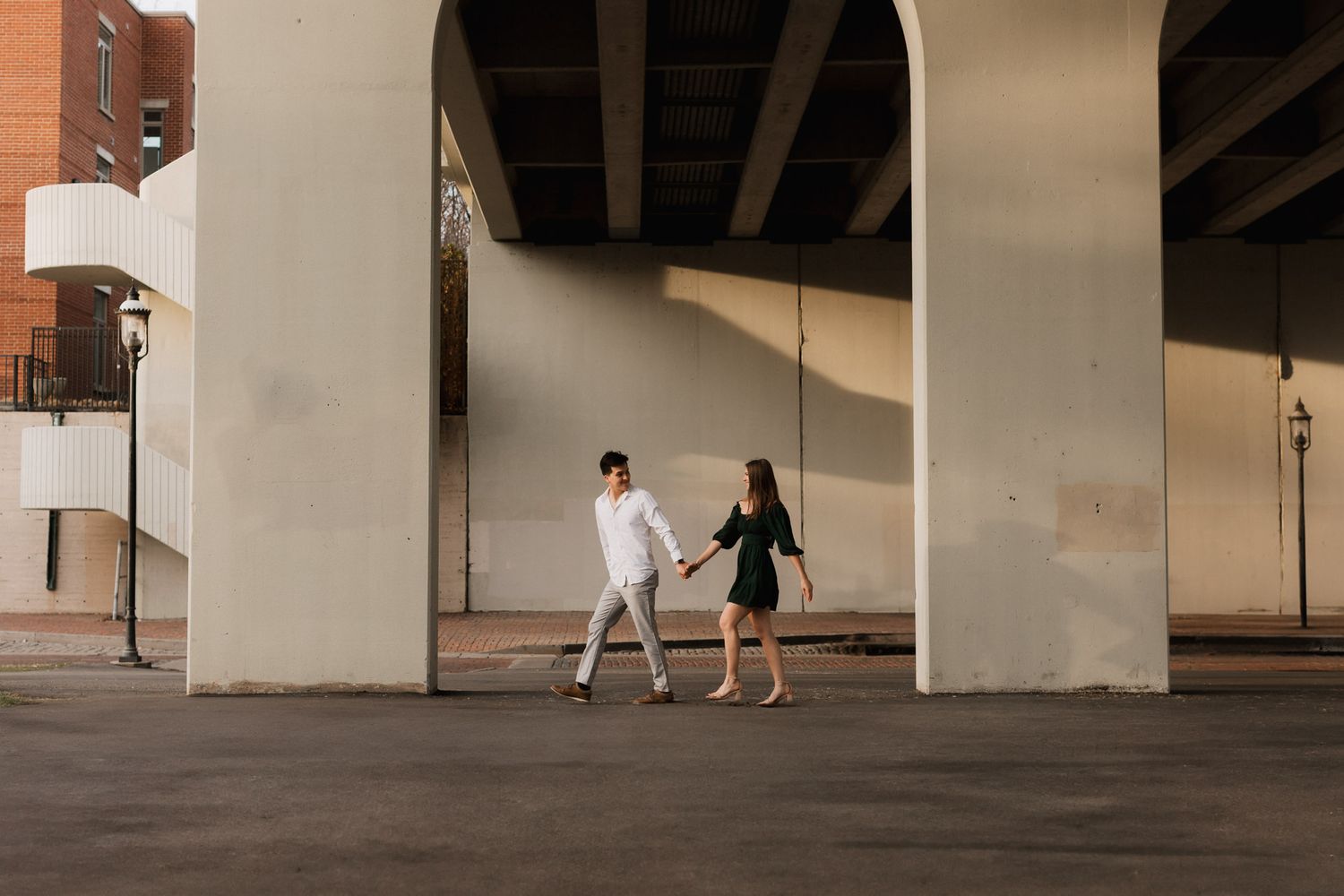 A couple holds hands walking under a large concrete bridge overpass in an urban setting.