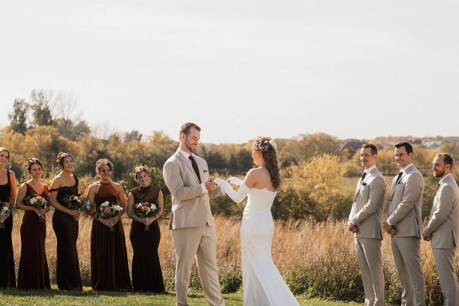 Bride and groom exchange vows outdoors in an autumn field as bridesmaids in rust dresses and groomsmen in tan suits look on.
