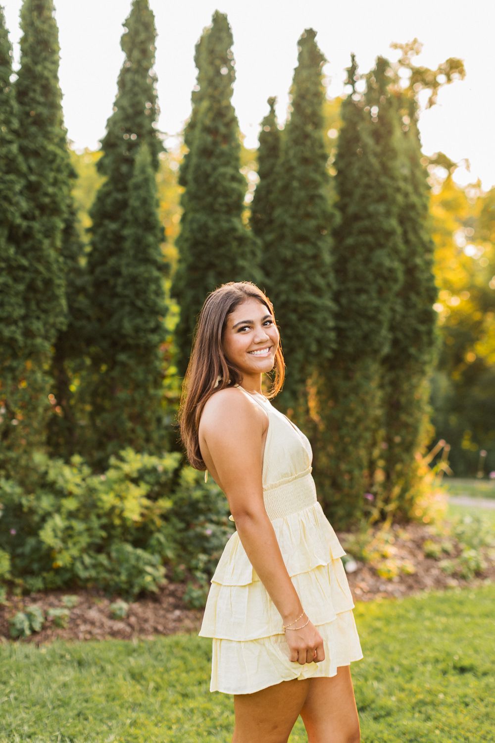 A person wearing a tiered yellow sundress poses in front of tall cypress trees during golden hour.