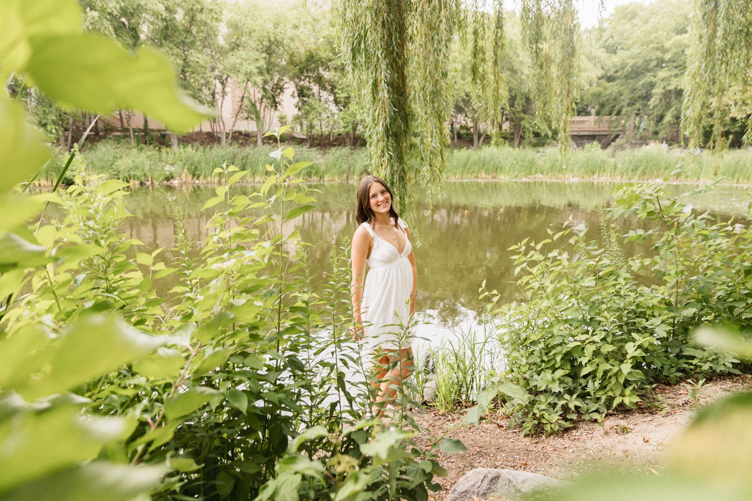 A dreamy outdoor scene with a person in a white dress standing by a serene pond surrounded by weeping willows and wildflowers.