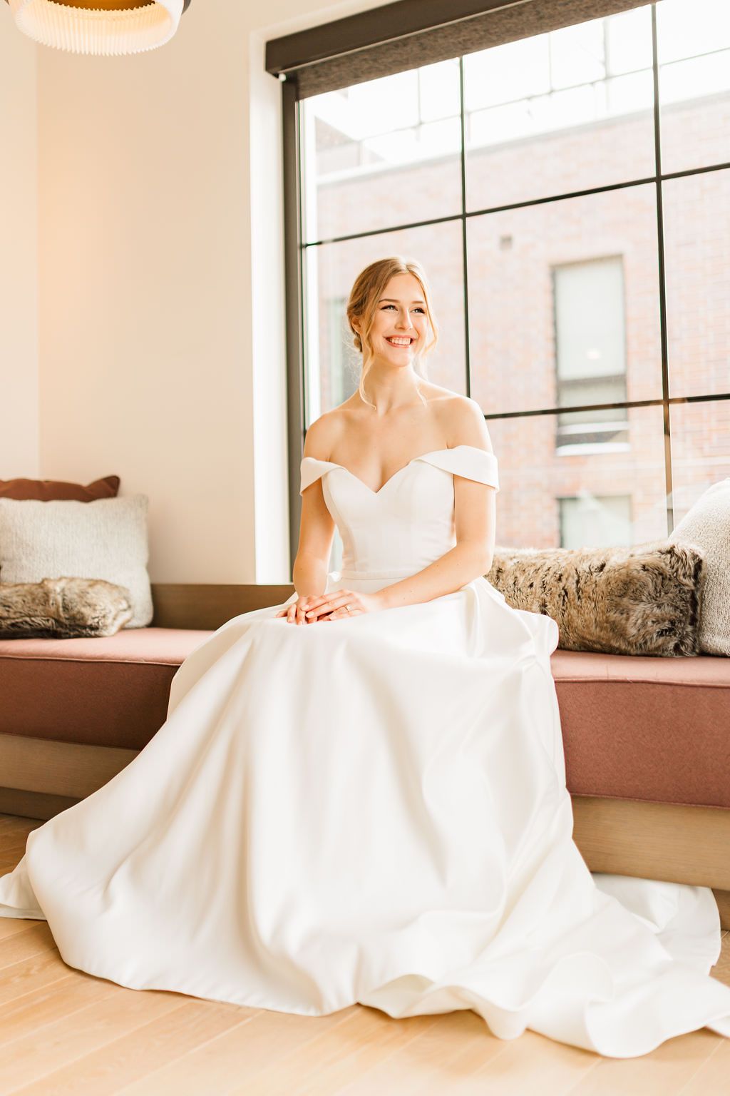 Light streams through windows as a bride in an elegant white gown sits on a bench in a minimalist room.