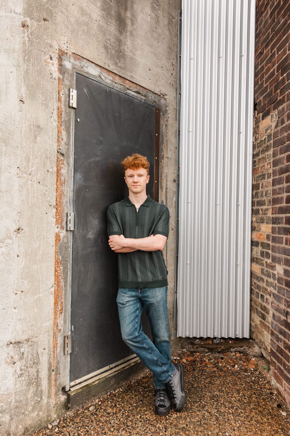 Figure poses against textured brick and concrete wall with corrugated metal elements in industrial setting.