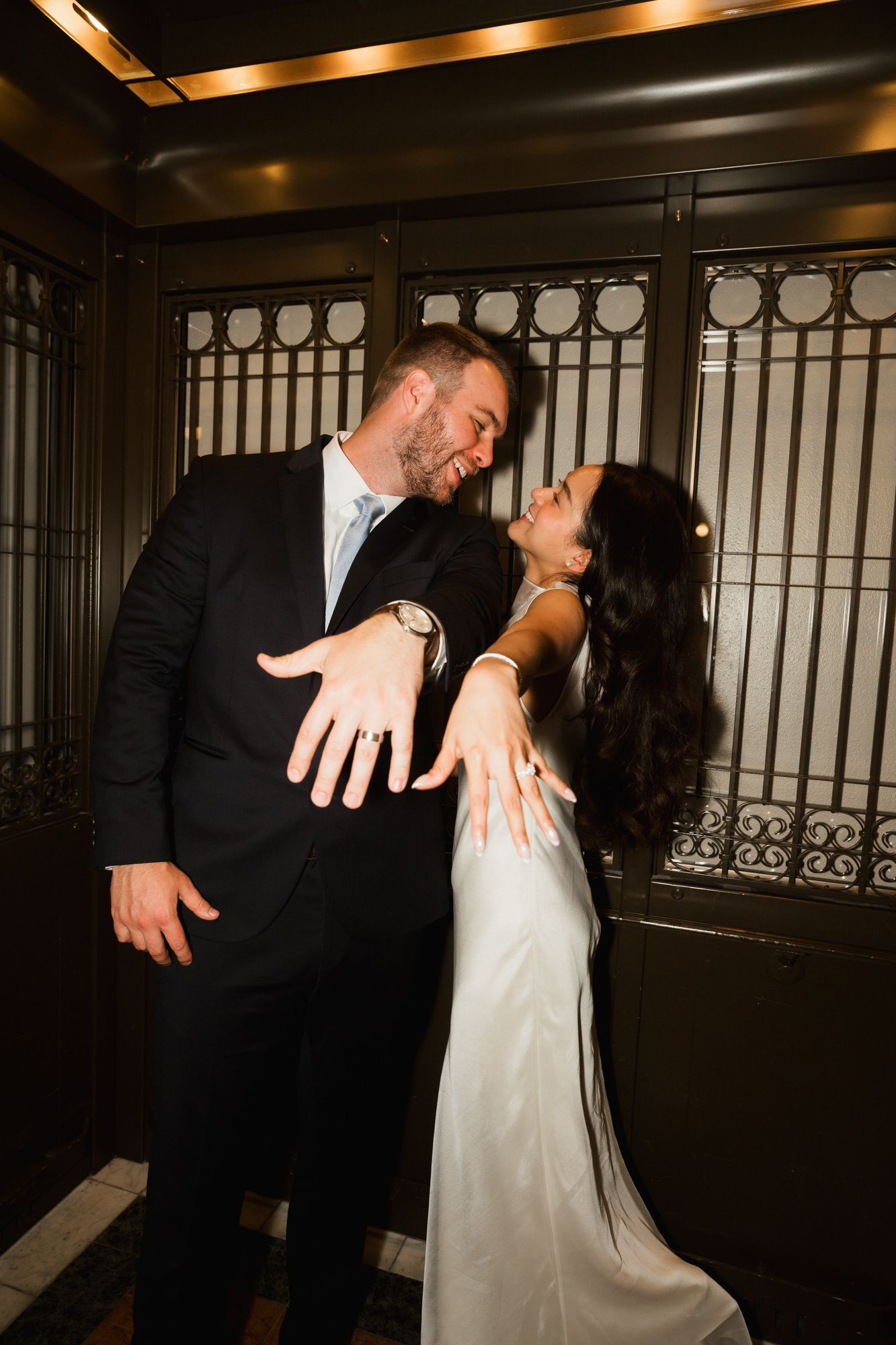 Newlywed couple sharing a romantic kiss, bride in white gown showing off wedding ring, groom in black suit.