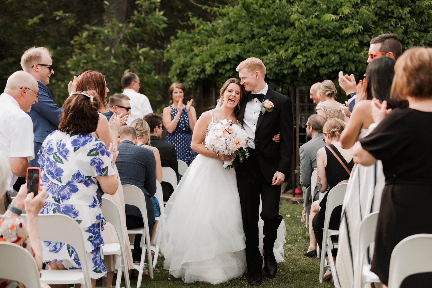 Bride and groom walk back down the aisle at outdoor wedding ceremony, guests applauding, surrounded by lush greenery.