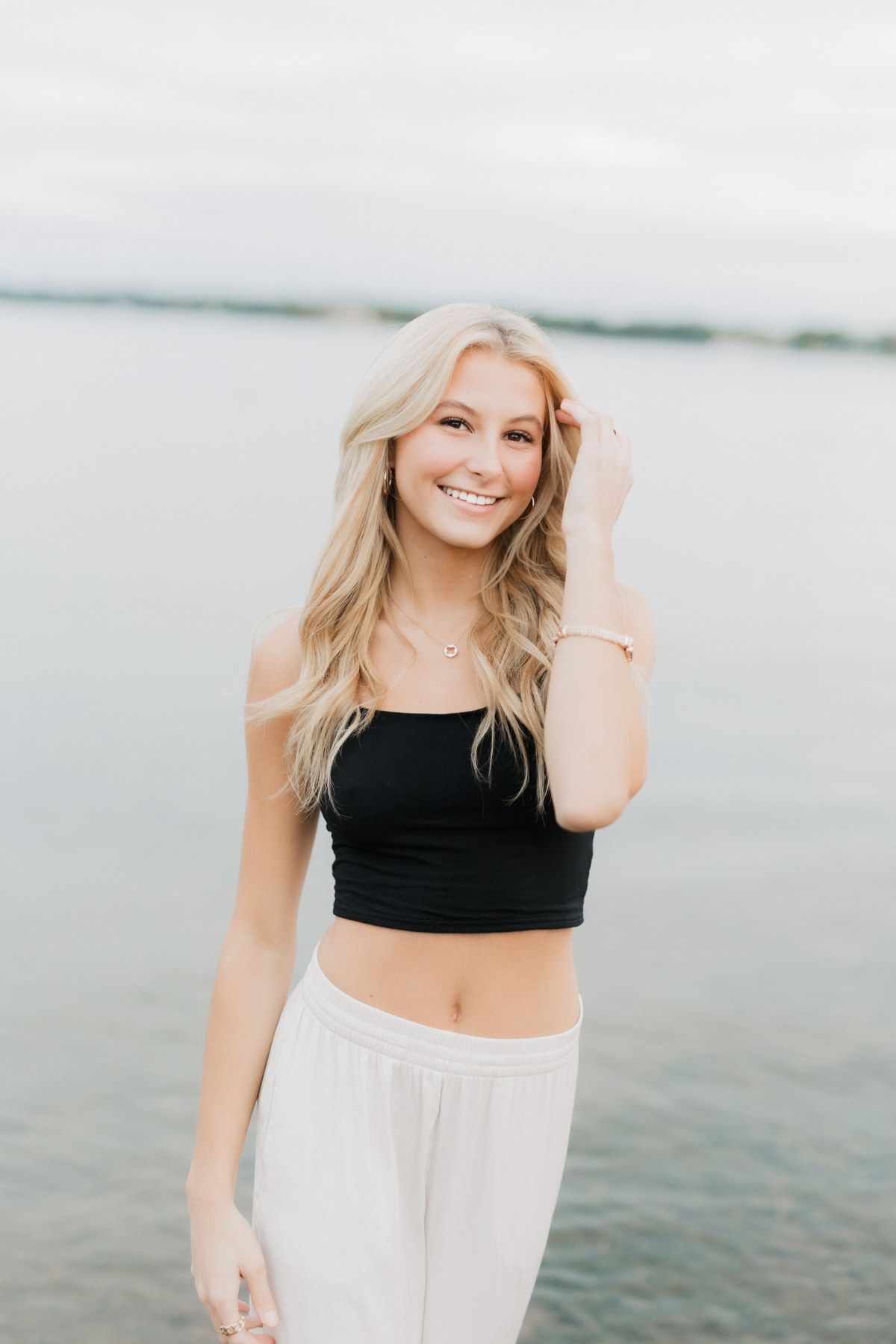 Smiling blonde woman in black crop top and white pants standing at lakeside beach.