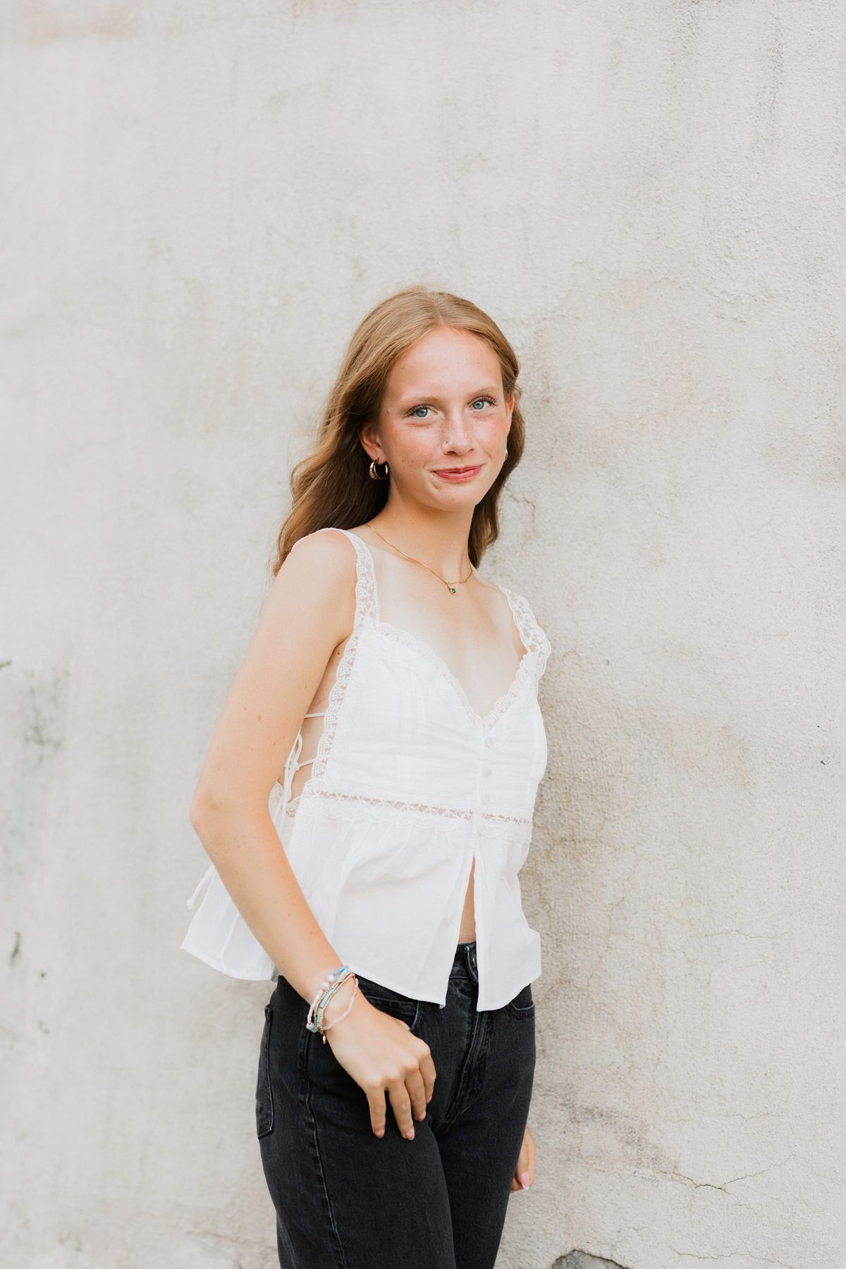 Young woman in white lace camisole and black jeans posing against a light concrete wall.
