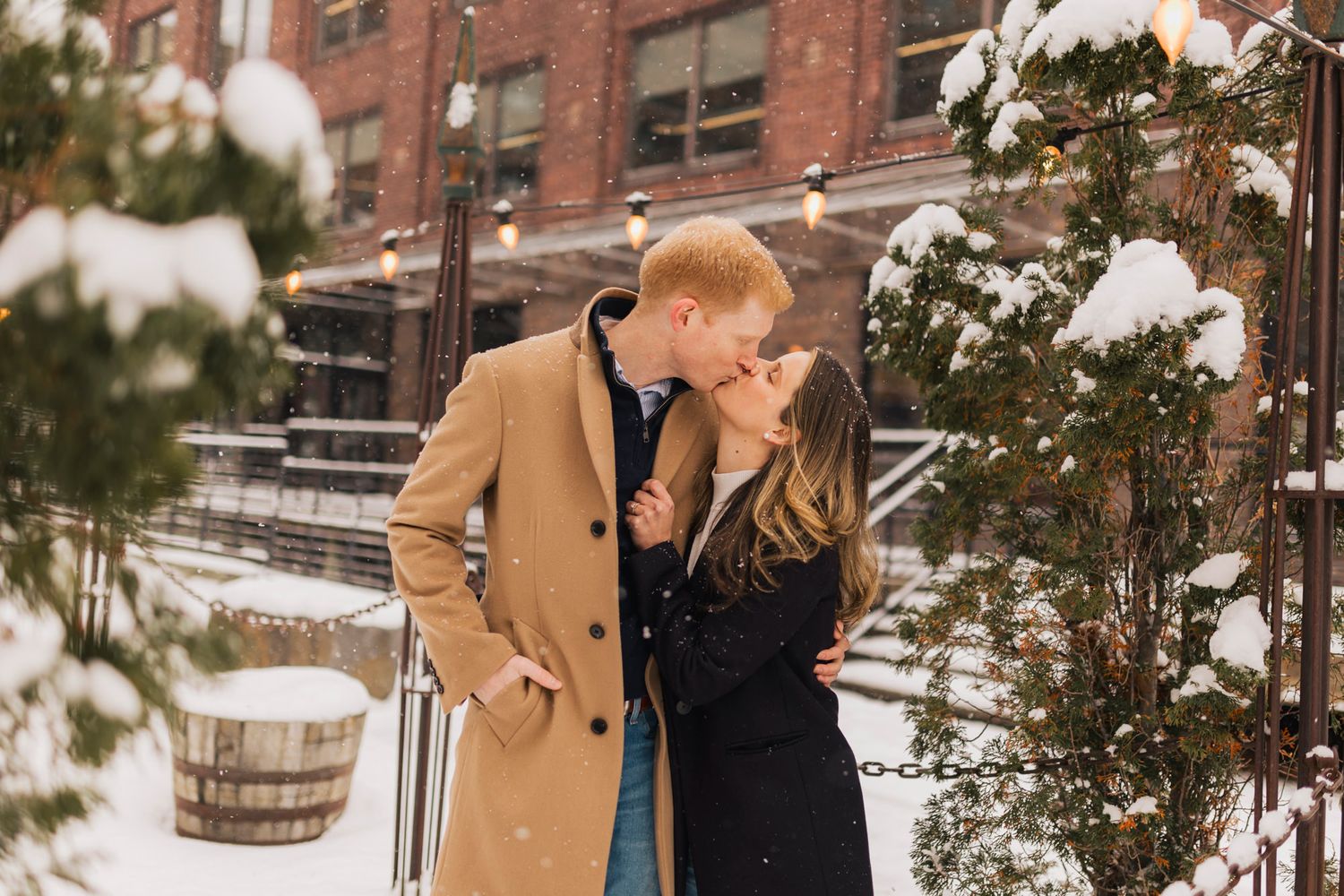 A romantic winter scene with a couple sharing a tender moment in front of a snowy brick building.