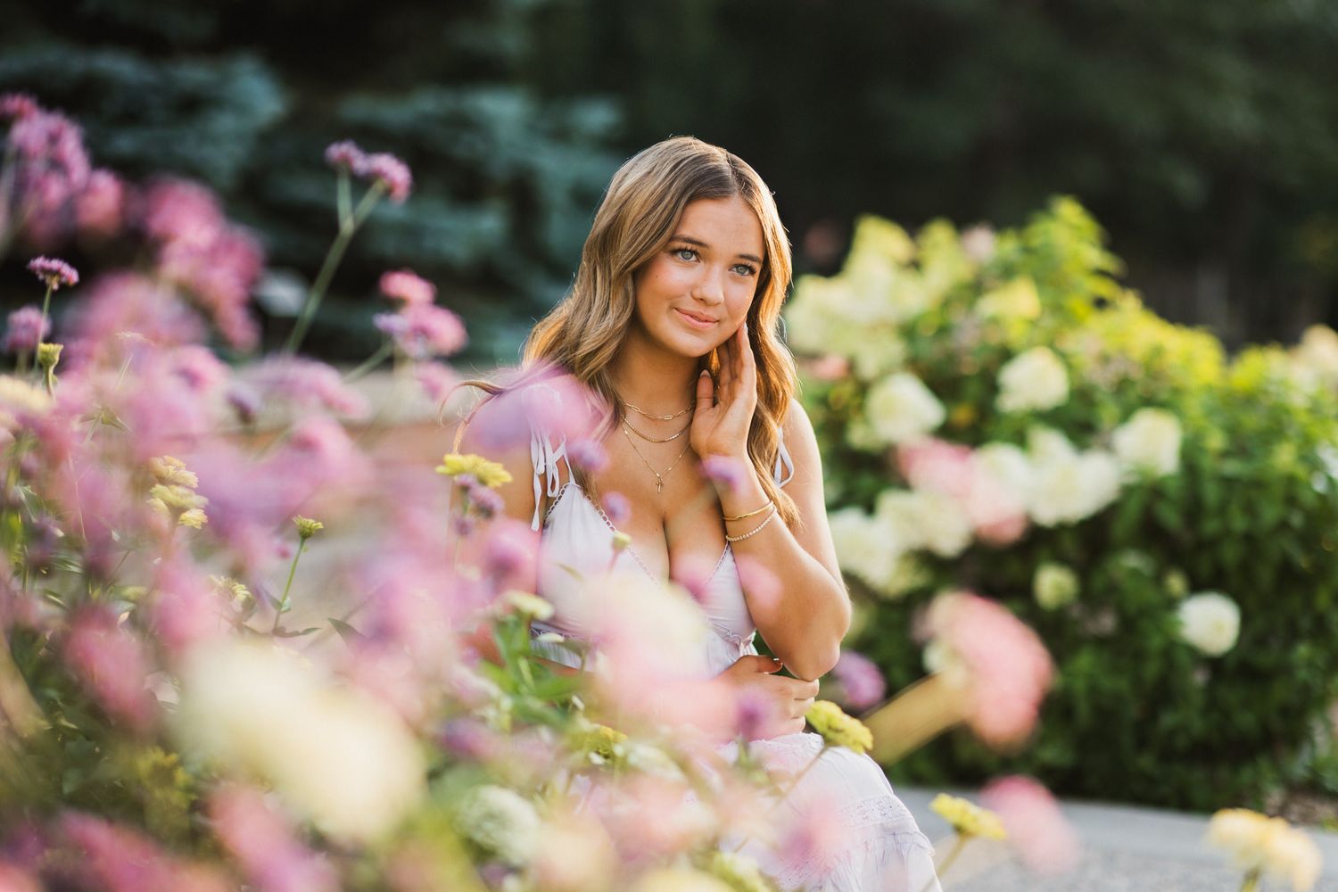 Young woman in floral dress sitting among pink and white garden flowers, smiling softly in a lush outdoor setting.