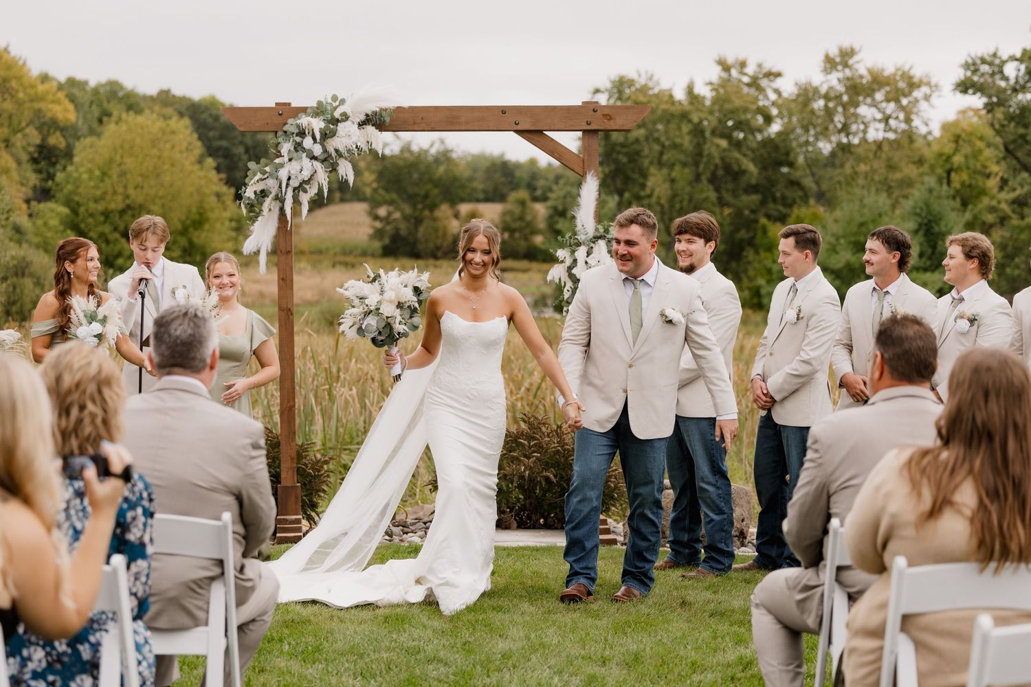 Bride and groom walk down the aisle at an outdoor wedding ceremony surrounded by guests and a wooden arch.