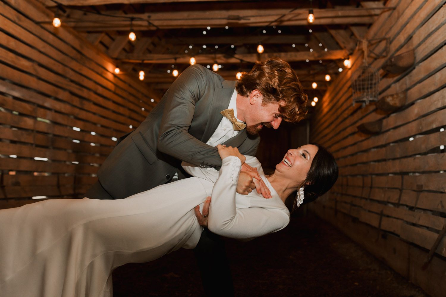 A romantic wedding couple dips and dances in a rustic wooden barn decorated with twinkling string lights.