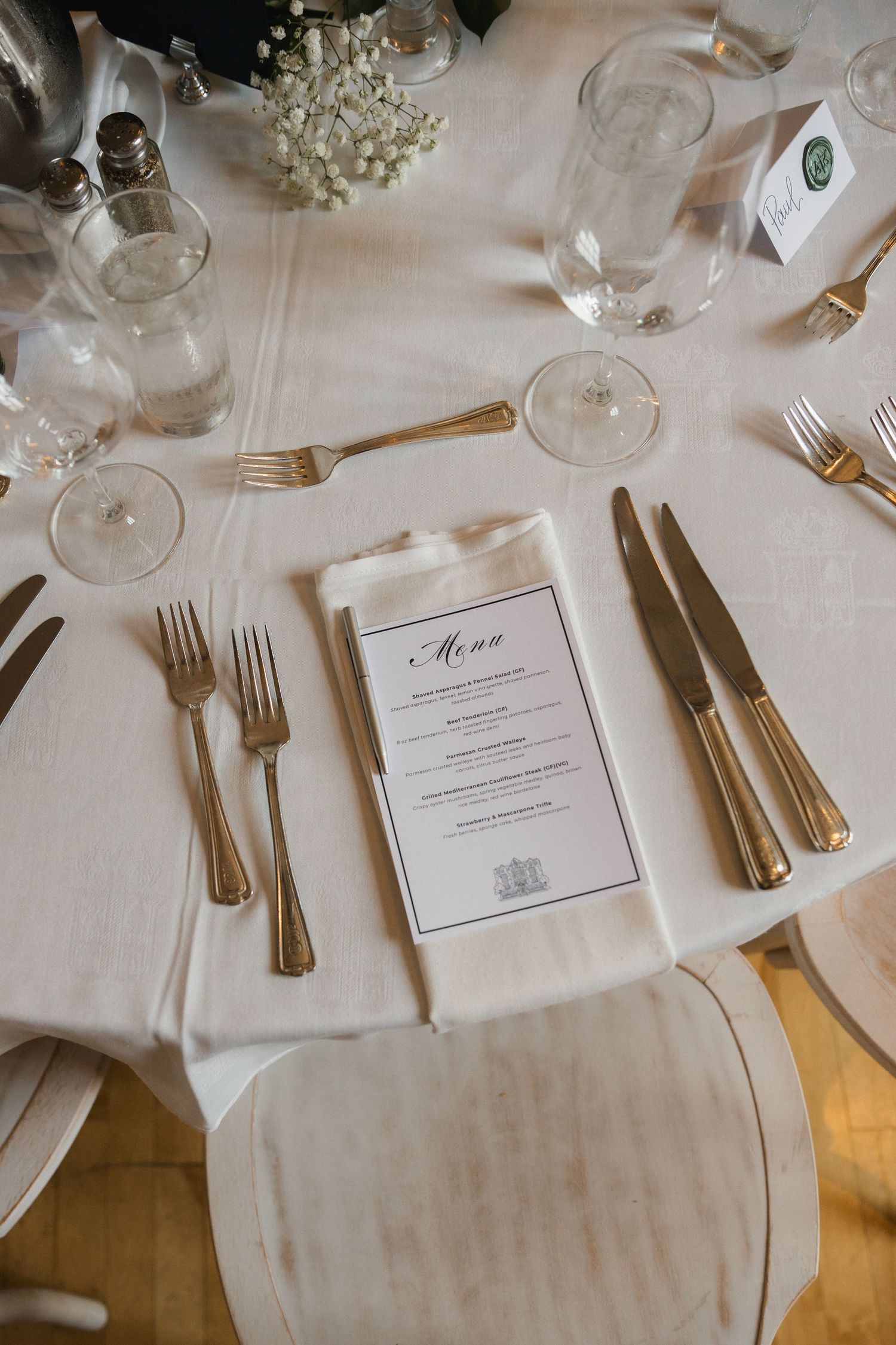 Elegant wedding reception place setting with gold cutlery, crystal glasses, and printed menu on white linen.
