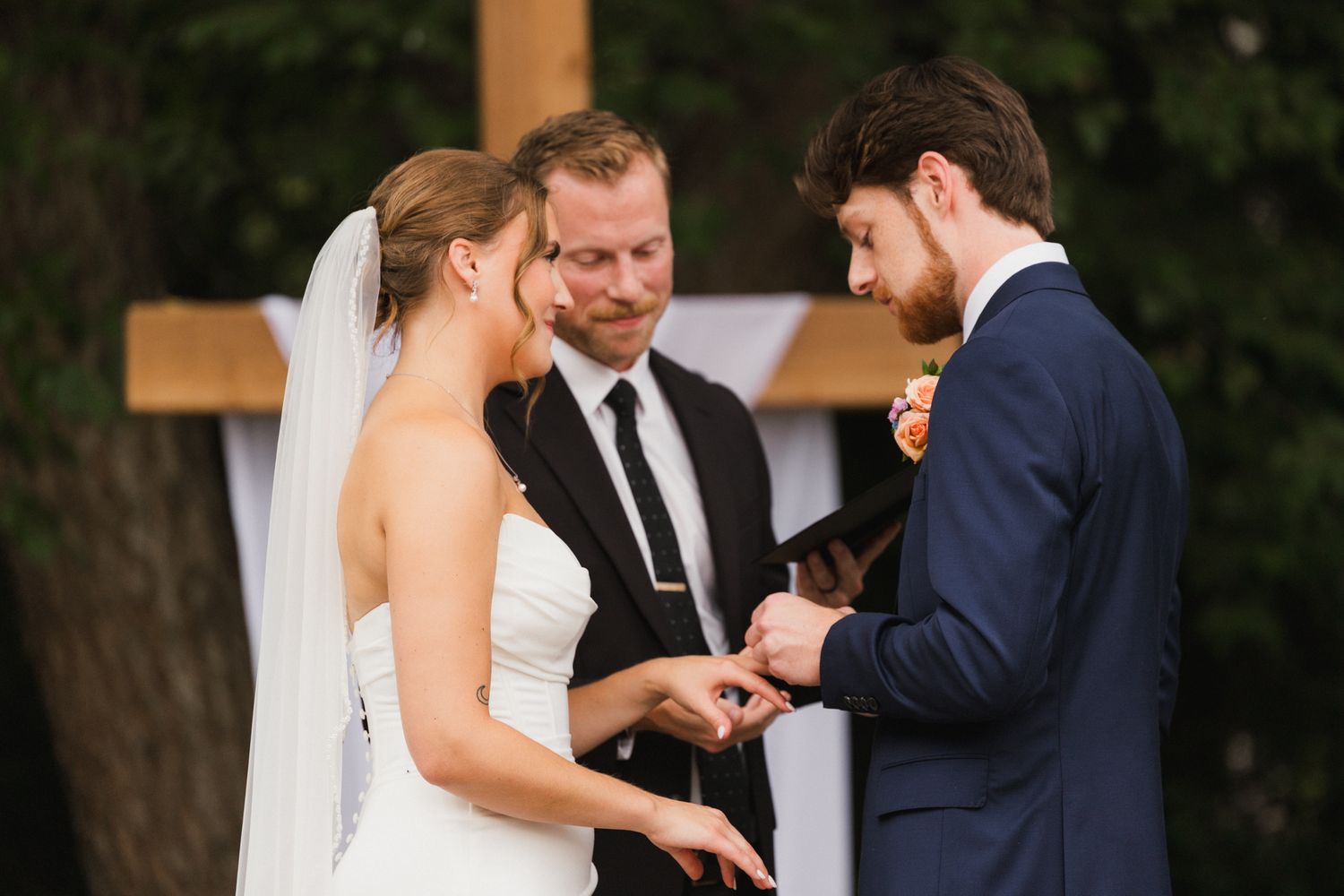 Groom placing wedding ring on bride's finger during outdoor ceremony with officiant and wooden cross backdrop.