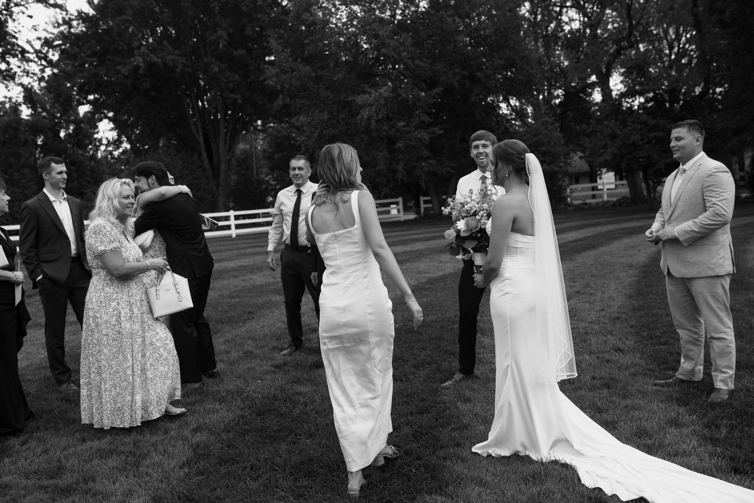 Black and white photo of a bride in white gown greeting wedding guests on a green lawn surrounded by trees.