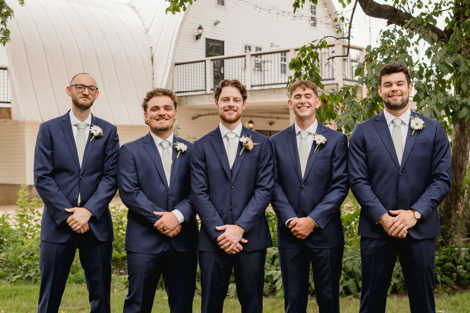 Five groomsmen in navy blue suits with floral boutonnieres posing outdoors at a wedding venue.