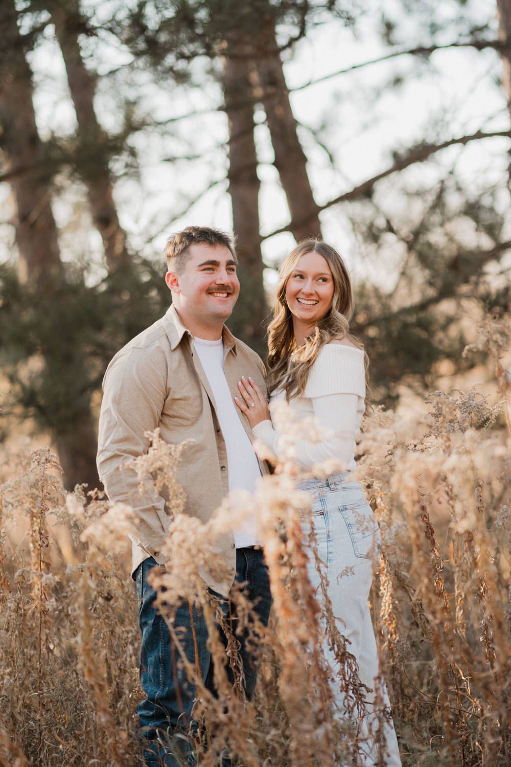 A couple laughs together in a field of dried grass with evergreen trees in the background.