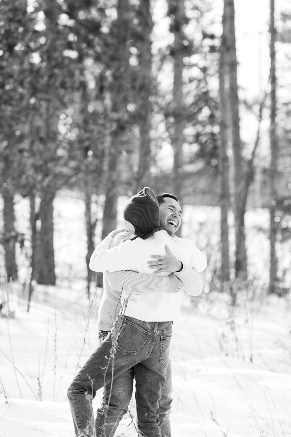 Two people embrace in a snowy winter forest setting wearing cozy winter clothing.