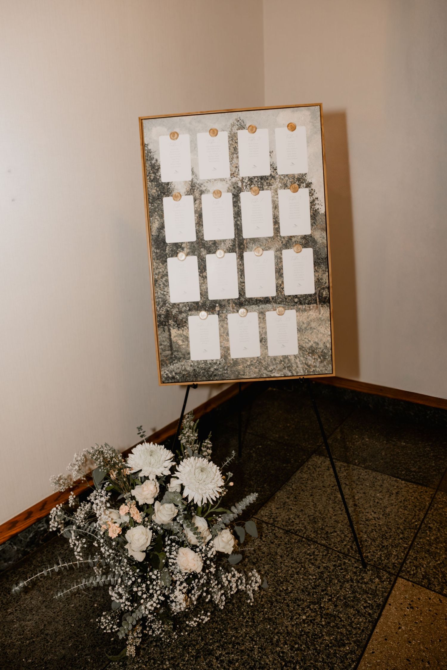 Elegant wedding seating chart on easel with white cards and gold accents, beside white floral arrangement.