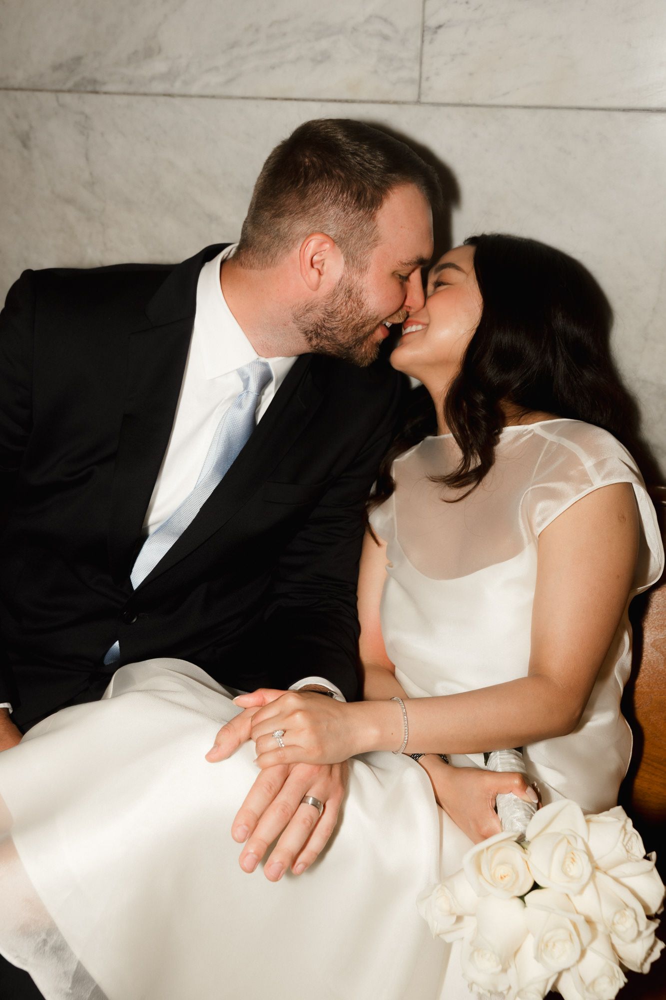 A bride and groom share a romantic kiss while seated, wearing wedding attire with a white bouquet in hand.
