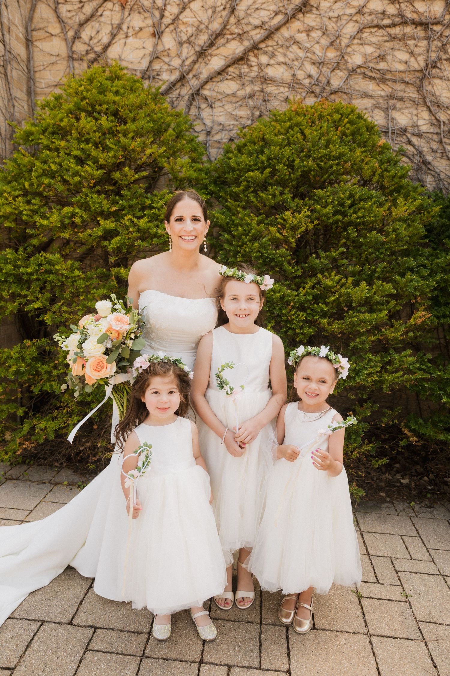 Bride in white strapless gown holds peach bouquet with three flower girls in white dresses and floral crowns outdoors.