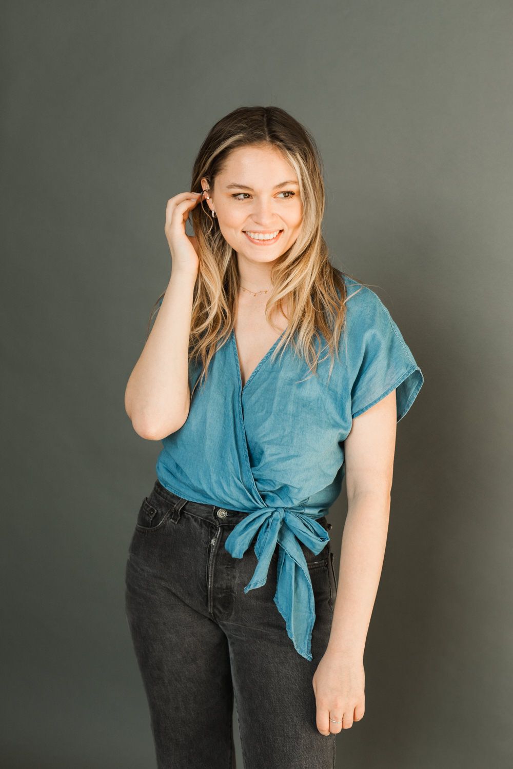 Young woman smiling in a teal wrap top and black jeans, posing against a dark gray background.