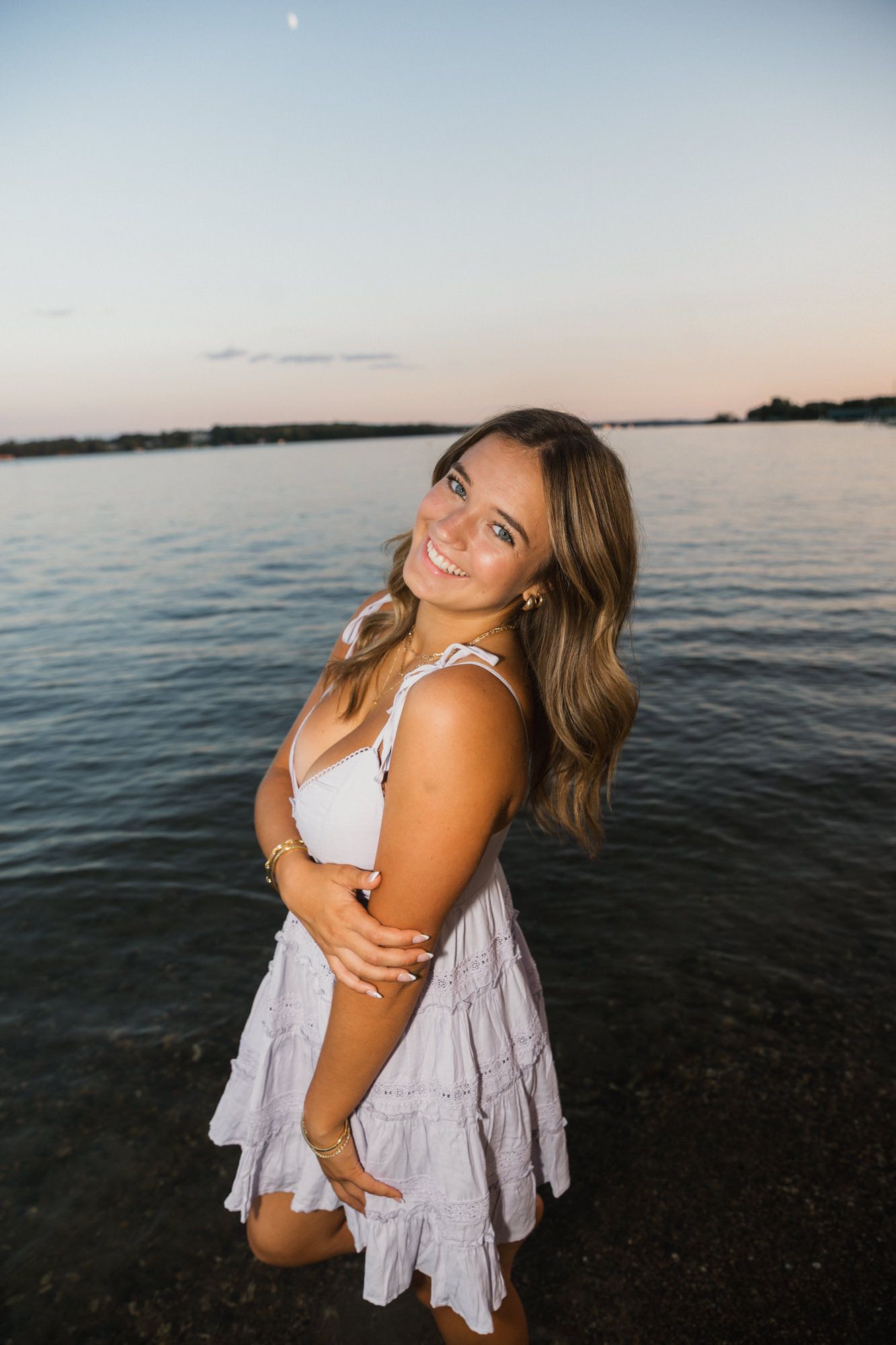 Young woman in white dress smiling by a lake at sunset, posing gracefully with flowing hair.